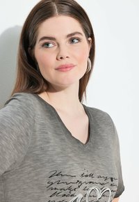 Šedé tričko s výstřihem do V, s texturovaným povrchem, krátkými rukávy a černým grafickým potiskem obsahujícím ručně psaný text a design srdce.