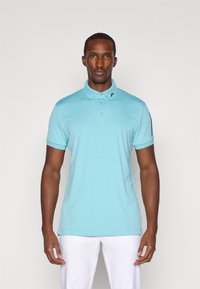 Lys blå polo skjorte i pustende materiale, med krage, tre-knappe placket, og en merket aksent på venstre skulder.