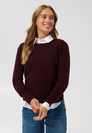 Ung kvinde med langt lyst brunt hår iført en hvid skjorte med krave under en mørk burgunder sweater og blå jeans, smilende og kigger mod højre.