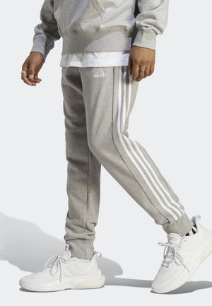 Pantalon de survêtement gris en tissu doux avec trois bandes blanches le long des côtés. Associé à des chaussures de sport blanches avec un design texturé.