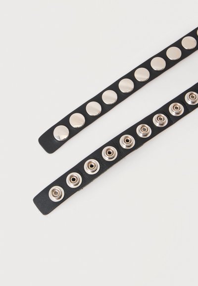 Deux sangles en cuir noir posées à plat, chacune avec une rangée de boutons-pression métalliques brillants pour la fermeture, présentées sur un fond clair.