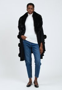Cappotto nero avvolgente con colletto in pelliccia sintetica, caratterizzato da dettagli a fiocco lungo l'orlo, indossato sopra un dolcevita bianco e jeans blu.