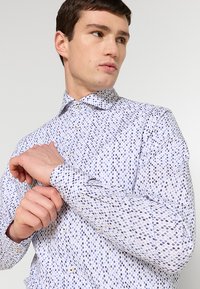 Witte shirt met een blauw geometrisch patroon, voorzien van een button-down kraag en lange mouwen. De stof lijkt glad met een lichte glans.