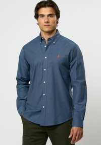 Man in een knopenblauwe lange mouwen shirt met een klein oranje logo op de borst, gecombineerd met donkergroene broek, staand met één hand in zijn zak.