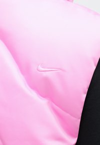 Pufasti prsluk izrađen od sjajnog ružičastog materijala s malim vezom bijelog Nike loga na prsima, u kombinaciji s crnom tkaninom ispod.