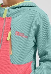 Giacca leggera con texture a nido d'ape, caratterizzata da un corpo turchese, un pannello rosa e dettagli verdi. Include un cappuccio e ricamo con il logo.