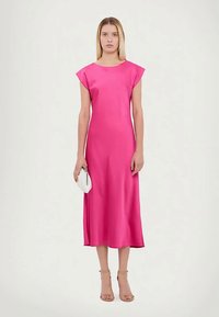 Ženska stoji v svetlo roza brezrokavnem midi obleku, drži majhno belo torbico, na nogah ima bež visoke pete, na enobarvni podlagi.