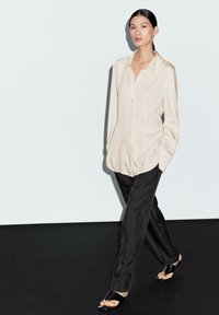 Camicia beige con bottoni, pantaloni neri a gamba larga, sandali neri a soffietto. Tessuto morbido, vestibilità rilassata, maniche lunghe, decorazioni minime.
