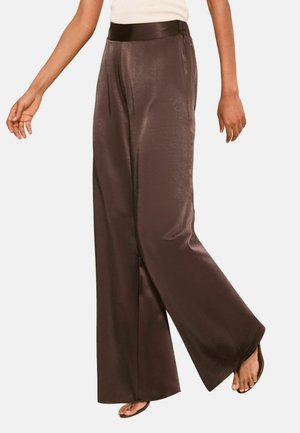 Žena nosí lesklé tmavohnedé široké nohavice s čiernym pásom, v kombinácii s otvorenými sandálmi, skrátené v páse a pri nohách.