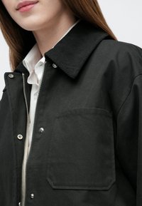 Geacă exterioară neagră cu guler texturat, fermoar argintiu, capse și buzunar pe pieptul stâng. Finisaj neted al țesăturii.