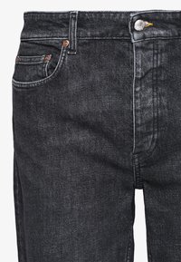 Primer plano de unos vaqueros de denim negro que muestran el bolsillo delantero, los lazos para el cinturón, el botón y los remaches de metal sobre una tela texturizada con un sutil desgaste.