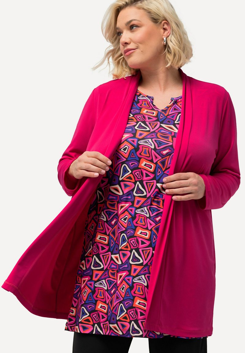 Fuchsia-Cardigan über einem gemusterten Kleid mit bunten, winkligen Designs in den Farben Lila, Rot und Weiß, mit einem V-Ausschnitt.