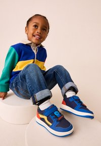 Baskets bleues et rouges avec une fermeture à scratch, tige texturée et un motif visage souriant. Semelle blanche avec un détail de bande jaune contrastante.