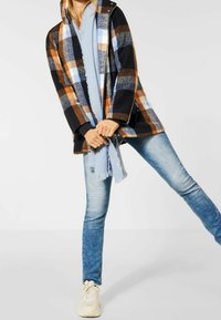 Svart och orange rutigt ytterplagg med huva, kombinerat med en ljusblå scarf, jeans av denim och beiga sneakers; synlig textur och mönster.