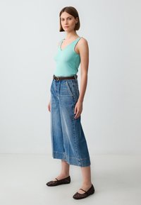 Un débardeur côtelé en bleu sarcelle clair associé à un jean en denim bleu clair à jambe large, avec une ceinture marron et des ballerines en mesh noir. Un design simple et décontracté.