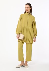 Olivgrüne, gefaltete Tunika mit hohem Kragen und Reißverschluss, kombiniert mit schmalen Hosen. Das Model hält eine kleine beigen Clutch und trägt weiße Mules.