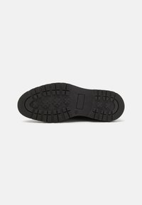 Semelle de chaussure en caoutchouc noir avec un motif de semelle profonde. Présente une texture antidérapante et un logo rectangulaire incrusté. Bout arrondi avec des bords renforcés.