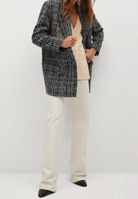 Blazer en tweed noir et blanc sur un pull crème à torsades, associé à un pantalon évasé crème et des escarpins noirs à bout pointu.