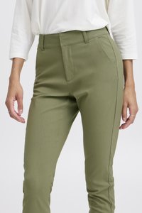 Femme portant un pantalon ajusté vert olive et un haut blanc à manches longues, debout les mains détendues sur les côtés sur un fond blanc.