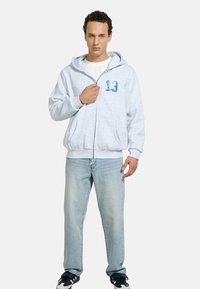 Young man standing, wearing light gray zip-up hoodie with blue number 13, white shirt, light blue jeans, and black and white sneakers.