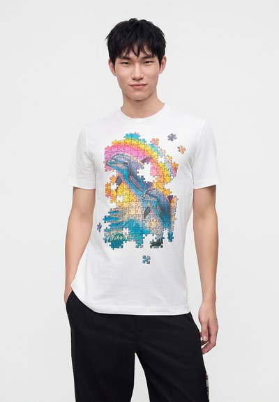 Νέος άνδρας που φοράει λευκό T-shirt με πολύχρωμο σχέδιο παζλ που δείχνει δύο δελφίνια και λείπουν σκορπισμένα κομμάτια παζλ.