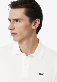 Bílá polo tričko vyrobené z bavlny s ribovaným límečkem, s vyšitým zeleným krokodýlím logem na hrudi. Čistý, minimalistický design.