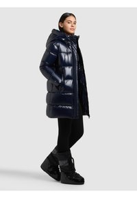 Sieviete tērpusies spīdīgā tumši zilā pūkainā mētelī ar kapuci, melnās legingās un melnās izolētās ziemas zābakos, stāv ar rokām kabatās.