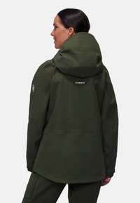 Giacca verde da esterno con un ampio cappuccio, polsini regolabili e un logo sul retro. Presenta una superficie liscia e resistente all'acqua.