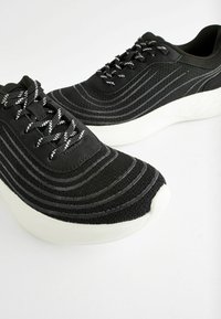 Black athletic shoes with textured fabric featuring horizontal stripes, complemented by white soles and contrasting black-and-white laces.
