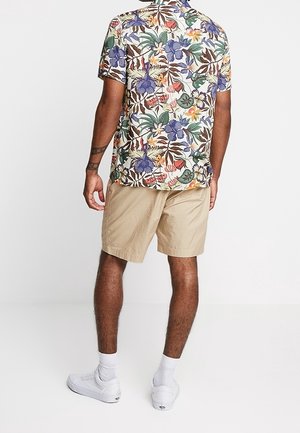 Hombre con pantalones cortos beige, zapatillas blancas y una camisa de manga corta con botones y un patrón floral colorido, de pie de espaldas.
