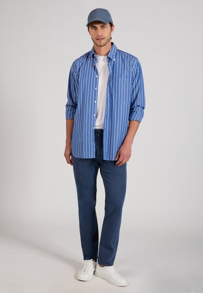 Man wearing blue striped button-up shirt over white t-shirt, blue pants, light blue cap, and white sneakers, standing against plain background.
