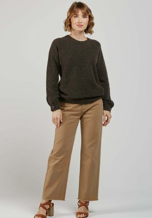 Femme aux cheveux courts portant un pull marron foncé, un pantalon à jambes larges beige et des sandales à talons à lanières marron, debout devant un fond uni.