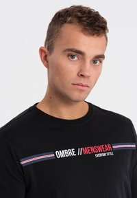 Schwarzes Baumwoll-T-Shirt mit rundem Halsausschnitt. Mit weißem und rotem Text "OMBRE // MENSWEAR EVERYDAY STYLE" und horizontalen blauen, roten und weißen Streifen.