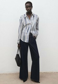 Prúžkovaná košeľa na gombíky v bielej a námorníckej modrej farbe, v kombinácii so širokými džínsovými nohavicami. Modelka drží malú čiernu tkanú kabelku. Hladká textúra, voľný strih.