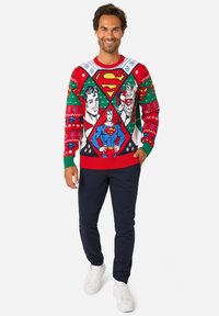 Červený pletený sveter s ilustráciami Supermana, zelenými a bielymi akcentmi a sviatočnými vzormi. Kombinovaný s tmavomodrými nohavicami a bielymi teniskami.