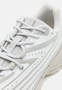 Leichtgewichtiger Laufschuh in Weiß und Hellgrau, mit einem strukturierten Obermaterial, aufwendigen Nähten und dezenten Perforationen für Atmungsaktivität.