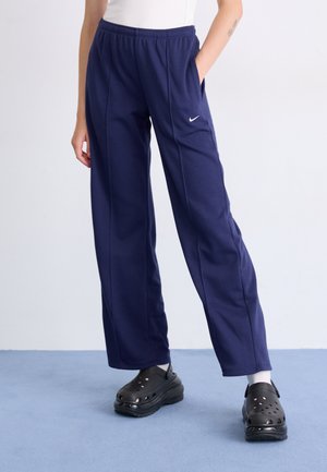 Person wearing navy blue Nike sweatpants, white socks, and black slip-on clogs standing on light blue floor.