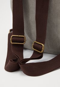 Sac en tissu gris avec des sangles en toile marron. Les sangles sont ornées de ferrures métalliques dorées. Gros plan mettant en valeur la texture et la forme.