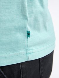 T-shirt in cotone blu chiaro con maniche corte, cuciture visibili e un piccolo etichetta del marchio color teal sul lato sinistro vicino all'orlo.