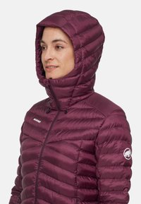 Fioletowa kurtka puchowa z poziomym wzorem pikowania, przypiętym kapturem, zapięciem na zamek błyskawiczny i logo na lewym rękawie. Gładki, błyszczący materiał.