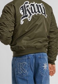 Olivově zelená bomber bunda s vyšitým černobílým "K" na zádech, spárovaná s modrými džíny s kontrastními designy kapes.