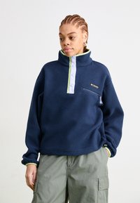Pullover in felpa blu navy con colletto alto, tasca con zip frontale e dettagli verde chiaro. Caratterizzato da patta con bottoni e vestibilità comoda.