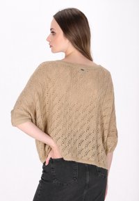 Pulover bej din tricot, cu un croi lejer, având un model de dantelă și mâneci scurte, rulate. Purtați cu blugi negri, pentru a evidenția o siluetă casual.