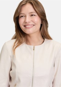 Jeune femme aux cheveux châtain clair souriant, portant une veste beige pâle à fermeture éclair devant un fond blanc.