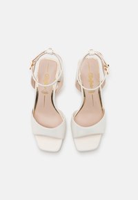 Vita satin klacksandaler med fyrkantig tå, ankelrem och guldaccente. Har en enkel design och vadderade beige innersulor.
