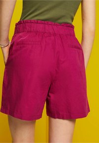 Shorts fucsias con cinturilla elástica, que cuentan con dos bolsillos traseros y un corte relajado. Textura suave sobre un fondo amarillo.