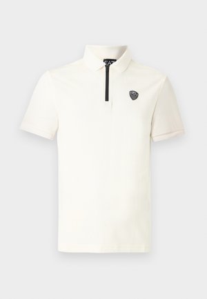 Lyhythihainen kreemunvärinen poolopaita, jossa on vetoketjullinen kaulus, litteä kaulus ja musta kilpi-logo vasemmassa rinnassa. Sileä puuvillakangas.