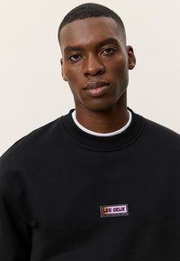 Νεαρός με κοντά μαλλιά που φοράει μαύρο φούτερ με διακριτικό λογότυπο "LES DEUX" στο στήθος, στέκεται μπροστά σε ανοιχτό φόντο.