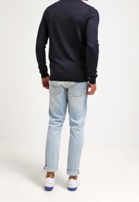 Pull bleu marine pour hommes en tissu tricoté, associé à un jean bleu clair. Le jean est légèrement usé, avec des revers roulés. Baskets blanches avec des accents violets.