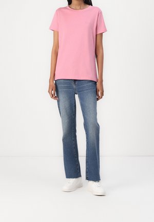 Person wearing a plain pink short-sleeve T-shirt, blue straight-leg jeans, and white sneakers standing against a white background.